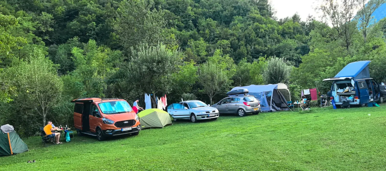 Kamp vozila, automobili i šatori poredani uz livadu, sa zelenim brdom u pozadini