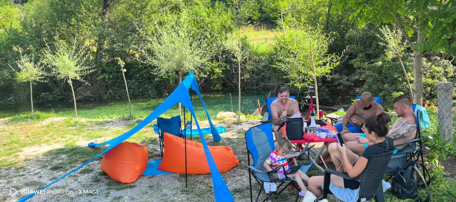 Gosti u kamp stolicama pod plavom tendom uz obalu Bijele, sa tirkiznom vodom u pozadini