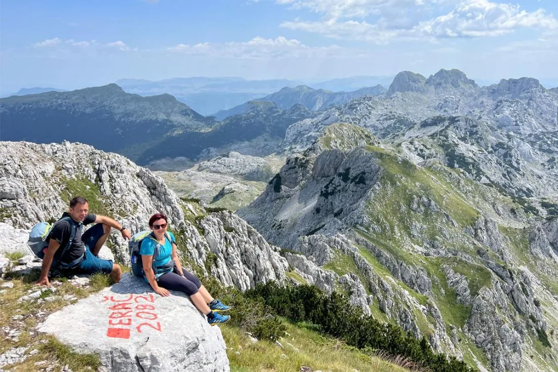 Dvoje planinara na vrhu iznad kamenog masiva Prenja, sa pogledom na okolne grebene
