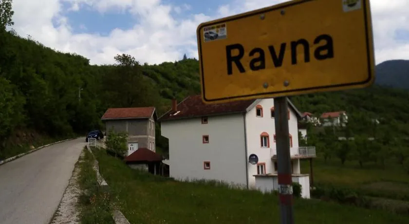 Žuta tabla sa natpisom Ravna na ulazu u selo, iza nje kuće i zelena brda